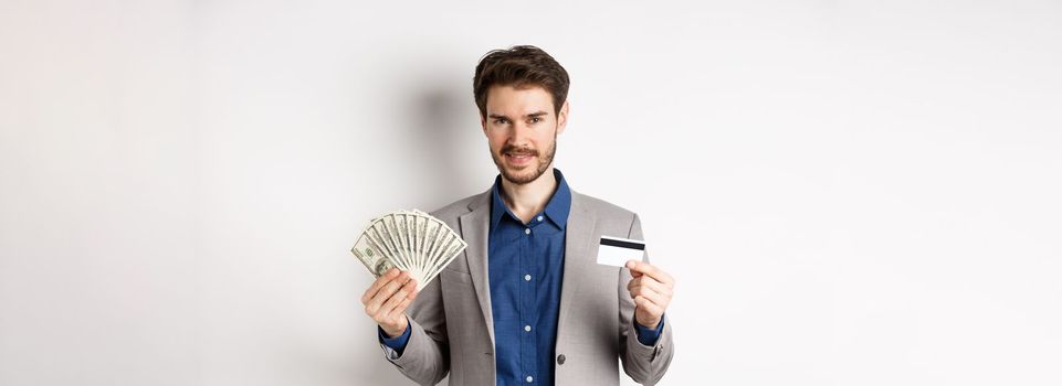 Rich smiling man in suit showing money and plastic credit card, standing with dollar bills and look pleased, white background.
