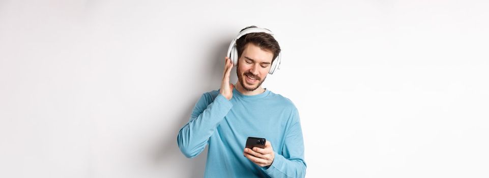 Cheerful smiling man listening music and looking at smartphone, reading message on phone, standing on white background.