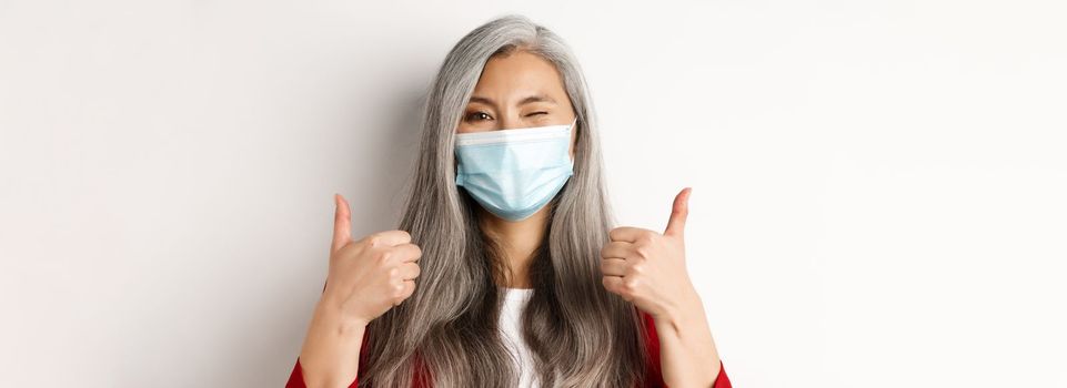Covid, pandemic and business concept. Cheerful asian businesswoman in medical mask, winking and showing thumbs-up in approval, white background.
