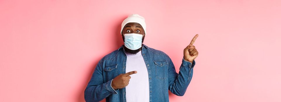 Coronavirus, lifestyle and global pandemic concept. Amazed black man in face mask showing advertisement, pointing at upper left corner and look excited, pink background.