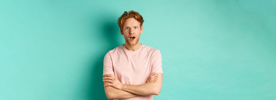 Intrigued redhead guy listening gossips, looking amazed and interested at camera, standing over turquoise background in casual t-shirt.