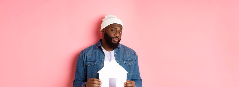 Real estate concept. Skeptical Black male model holding paper house model, looking suspicious and doubtful at camera, searching apartment, standing over pink background.
