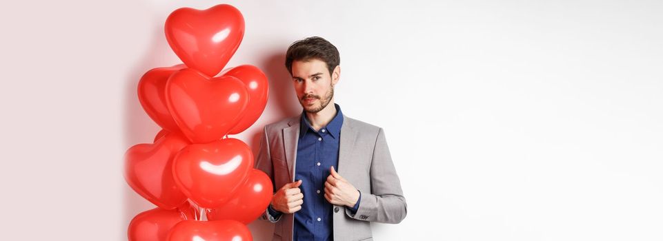 Lovers day. Handsome and confident young man getting dressed for Valentines day, fixing suit and looking at camera, standing near romantic heart balloons, white background.