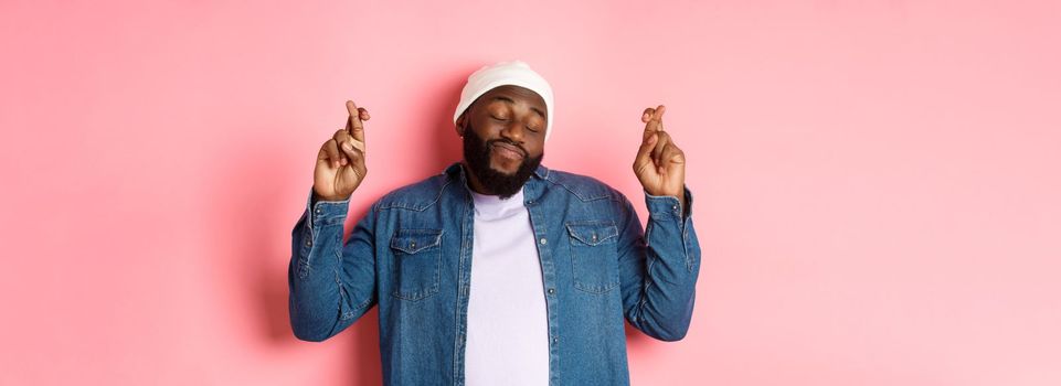 Hopeful and dreamy Black man making wish, cross fingers and smiling, standing over pink background.