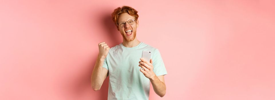 Happy redhead guy in glasses and t-shirt winning online prize, shouting yes with joy and satisfaction, holding smartphone and making fist pump, pink background.