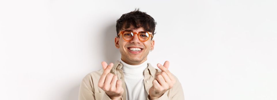 Cute young guy in glasses smiling and showing finger hearts, standing on white background.