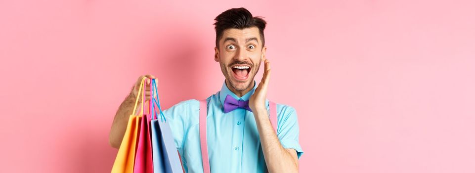 Surprised and happy man screaming of joy and holding shopping bags, shopper seeing discounts, standing over pink background.