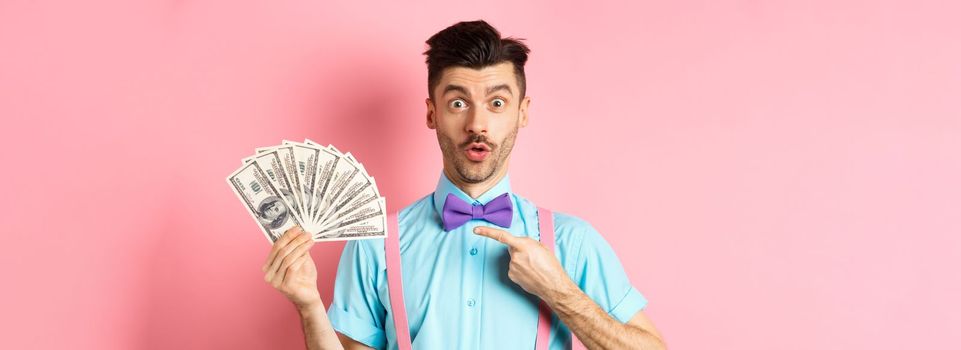 Amazed man showing big money prize, pointing at dollars and say wow, stare impressed at camera, standing over pink background.