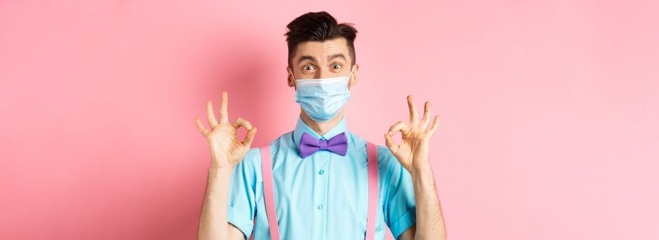 Coronavirus, healthcare and quarantine concept. Happy guy in medical mask and festive bow-tie showing all good gesture, make okay signs and smile at camera, pink background.