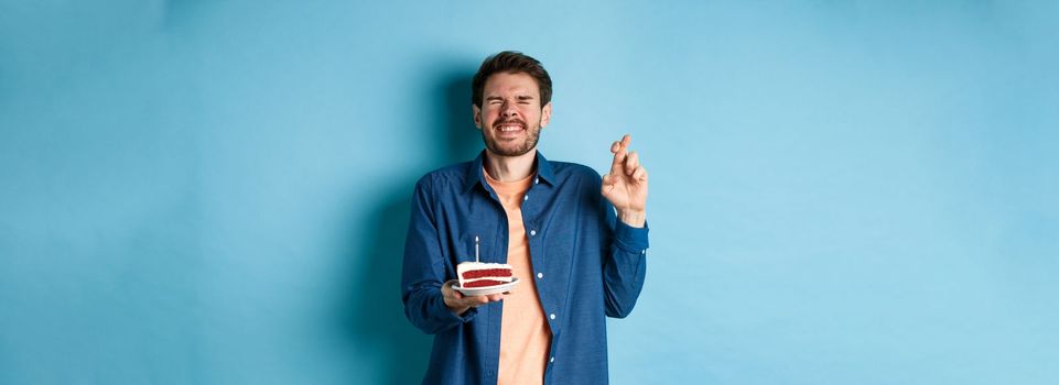 Celebration and holiday concept. Hopeful guy cross fingers and making birthday wish on cake candle, standing on blue background.
