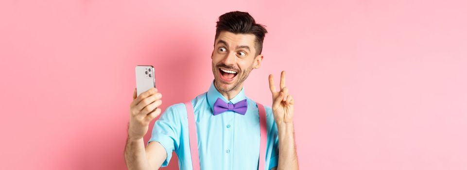 Technology concept. Funny man with moustache and bow-tie taking selfie on smartphone, showing peace sign and smiling at mobile camera, pink background.