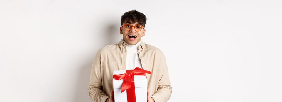 Surprised happy guy receiving a gift, holding present box and smiling amazed, standing on white background.