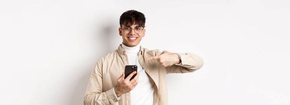 Real people. Handsome young man in glasses pointing finger at smartphone screen, showing online promo, standing on white background.