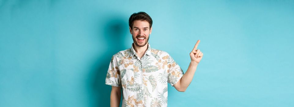 Cheerful young man in hawaiian shirt pointing finger left, showing empty space and smiling. Concept of tourism and vacation.