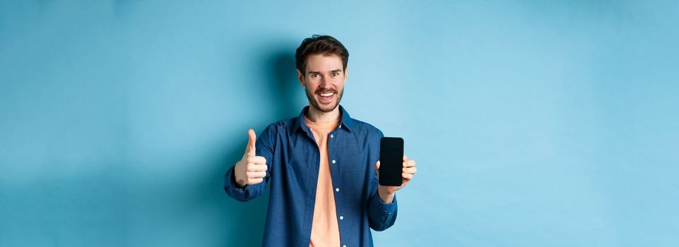 Handsome young man showing thumbs up gesture and empty smartphone screen, recommending app or company, standing on blue background.