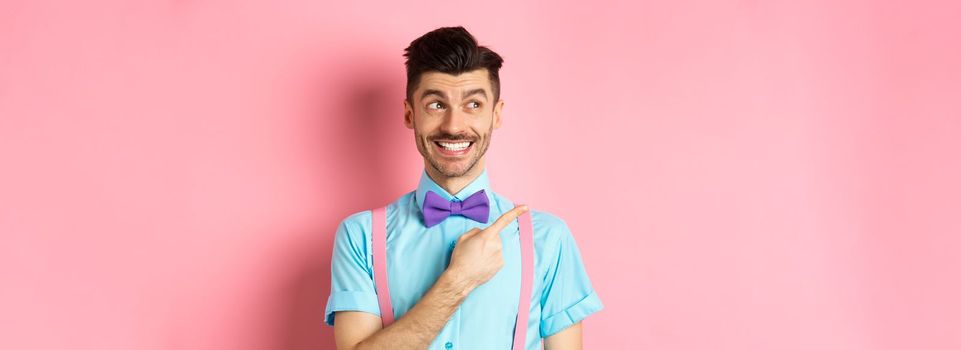 Happy smiling man in funny outfit and bow-tie, pointing finger left and looking at logo, showing advertisement on pink background.
