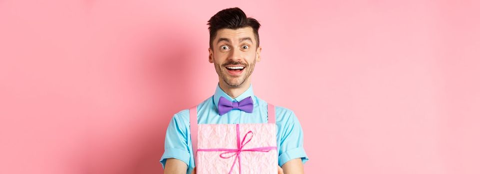 Holidays and celebration concept. Cheerful guy wishing happy birthday and giving you gift wrapped in box, standing over pink background in festive clothes.