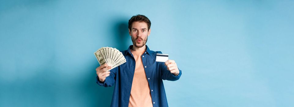 Excited modern guy showing money and plastic credit card, saying wow amazed, standing on blue background.
