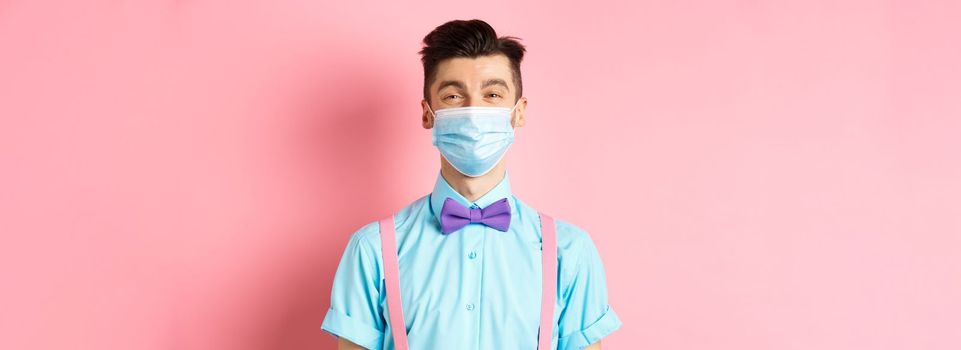 Covid-19, pandemic and health concept. Portrait of smiling man in medical mask feeling happy, standing in festive bow-tie and suspenders, pink background.