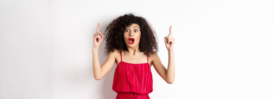 Impressed young woman with curly hair, wearing red dress, gasping and saying wow, pointing fingers up at logo, standing over white background.