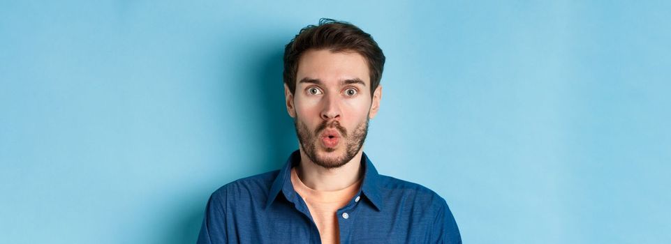 Close up portrait of amazed and surprised man saying wow, staring impressed at camera, checking out cool offer, standing on blue background.