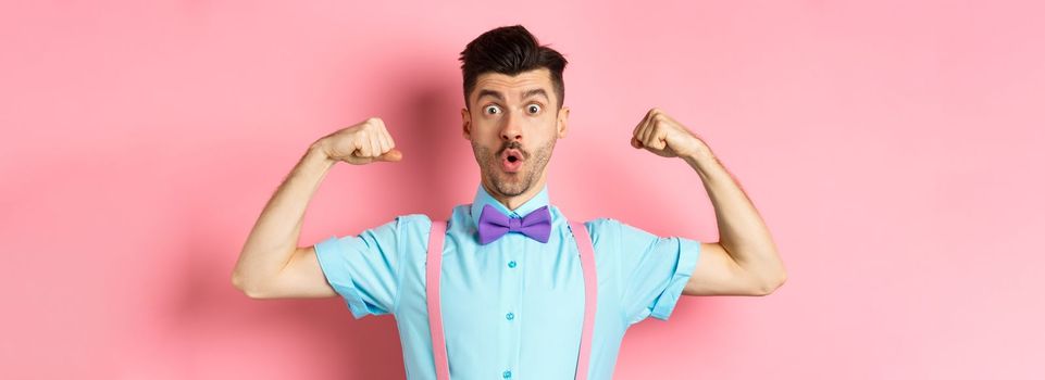 Image of funny guy with moustache and bow-tie, showing muscles, flexing biceps and looking surprised at camera, standing in disbelief on pink background.