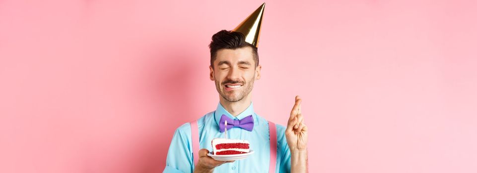 Holidays and celebration concept. Hopeful birthday guy in party hat making wish, holding cake with candle and cross fingers for dream come true, pink background.