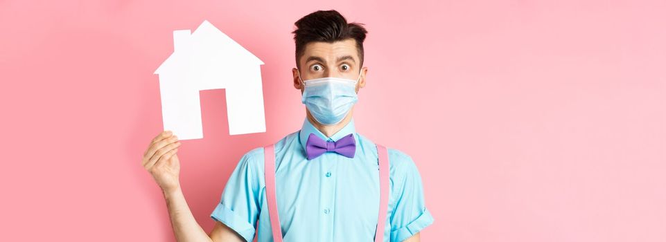 Covid, pandemic and real estate concept. Confused guy in medical mask searching property, showing house cutout and stare at camera worried, standing over pink background.