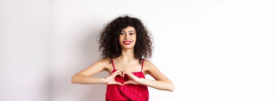 Charming young woman with red romantic fress, showing heart gesture and smiling, express love, standing over white background.