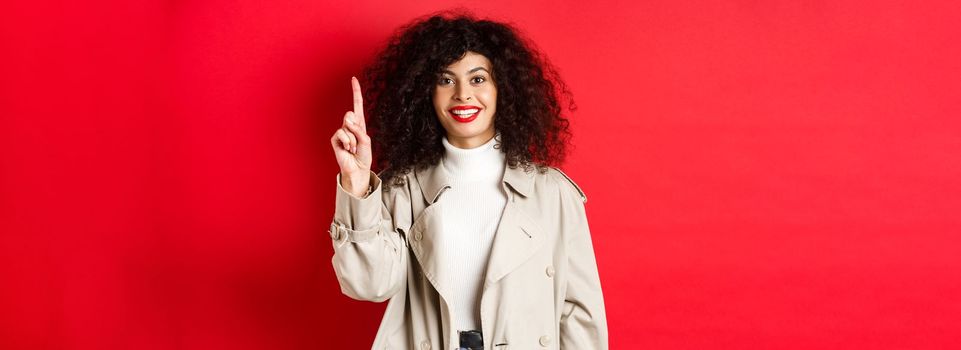 Attractive modern woman with red lips, curly hairstyle, wearing spring trench coat, pointing finger up and smiling, standing against red background.