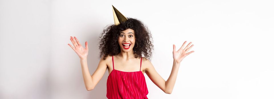 Cheerful young woman in red dress, celebrating birthday, wearing party hat and smiling, screaming of joy, standing on white background.