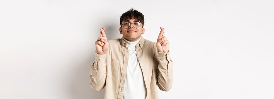 Hope. Happy young man making wish and smiling, looking hopeful at camera as waiting for dream come true, cross fingers for good luck, standing on white background.