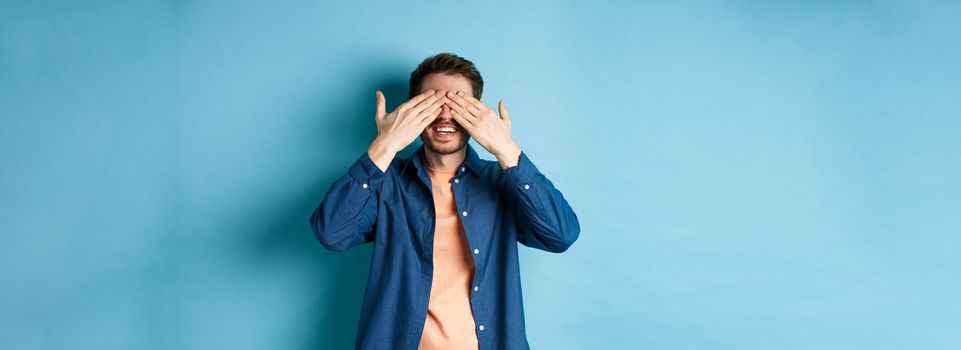 Happy smiling guy waiting for surprise, covering eyes with hands and anticipating gift, standing on blue background.