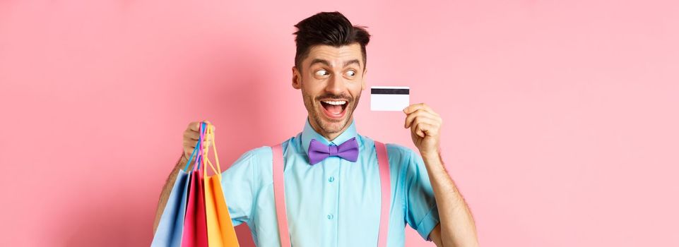 Cheerful funny guy looking at plastic credit card and smiling, buying gifts, holding shopping bags, standing on pink background.