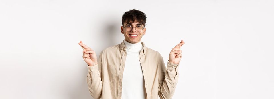 Handsome and positive young man smiling, making wish with crossed fingers and happy face, hope for dream to come true, standing on white background.