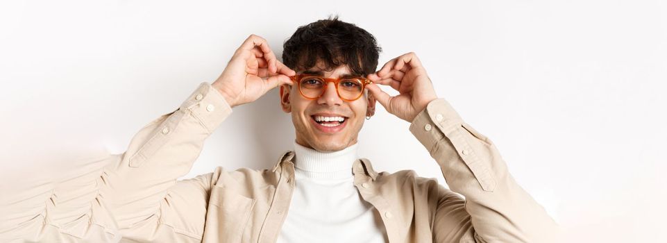 Close-up of stylish hipster guy trying eyewear at optician store, put on glasses and smiling, standing on white background.