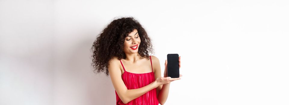 Beauty woman with makeup and curly hair, showing empty smartphone screen, demonstrate app, standing on white background.