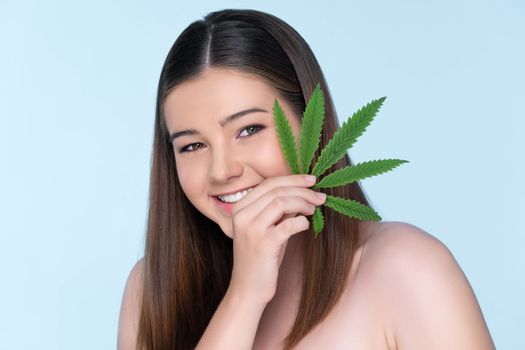 Closeup portrait of charming girl with fresh skin holding green leaf for beauty skin care made from cannabis leaf. Cosmetology and cannabis concept with isolated background.