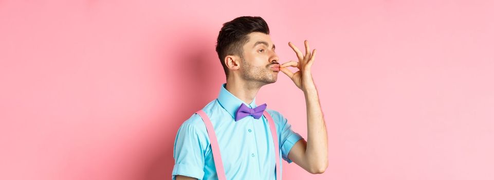 Profile of funny guy touching his french moustache and pucker lips like snob, standing smug on pink background and look left.
