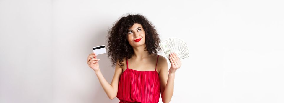 Shopping. Woman thinking and smiling, holding money with plastic credit card, wearing red elegant dress and evening makeup, white background.