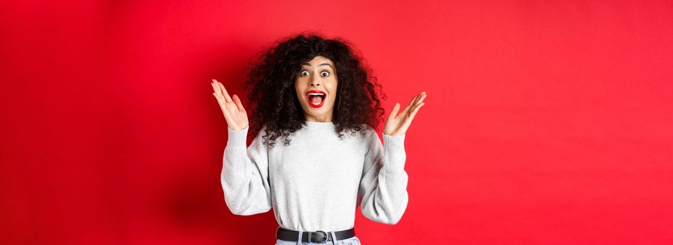 Image of happy and surprised curly woman in makeup and sweatshirt, raising hands up and rejoicing from good news, standing on red background.