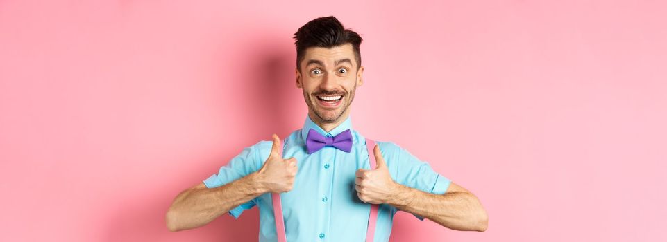 Happy young man showing thumbs up and smiling, saying yes, agree with something cool, recommending excellent deal, standing upbeat on pink background.