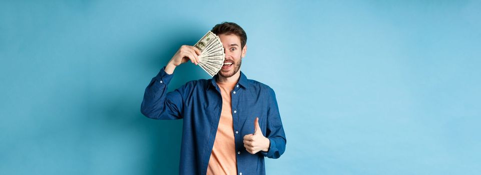 Cheerful guy cover half of face with money and showing thumbs-up, recommending fast cash loan, standing on blue background.