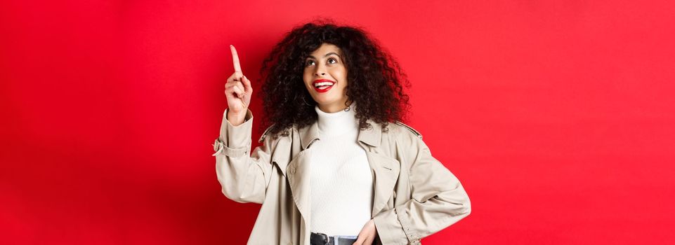 Excited woman with curly hair, wearing trench coat, pointing at upper right corner and smiling amazed, standing against red background.
