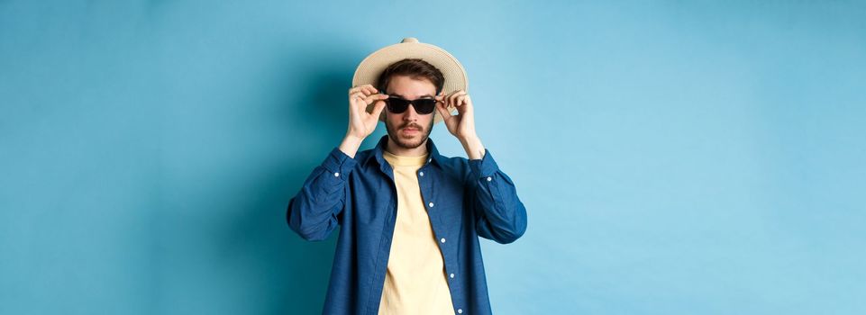 Handsome tourist in straw hat put on sunglasses on summer vacation, standing on blue background.