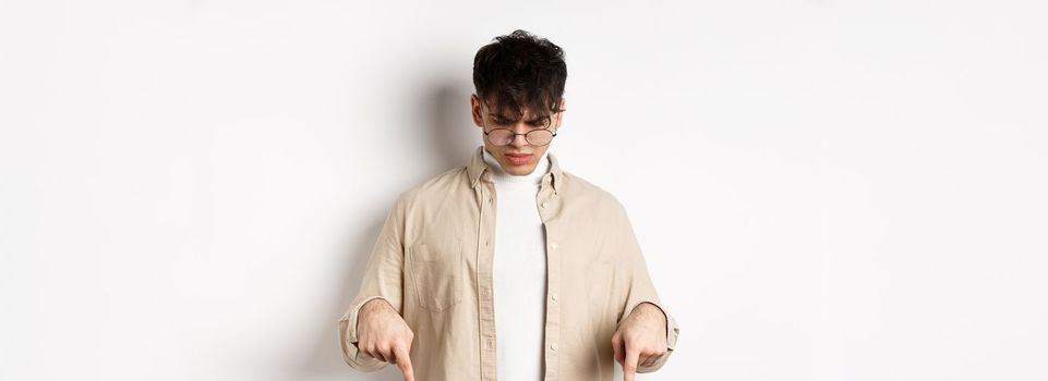 Confused frowning guy in glasses looking, pointing fingers down at something strange, standing puzzled on white background.