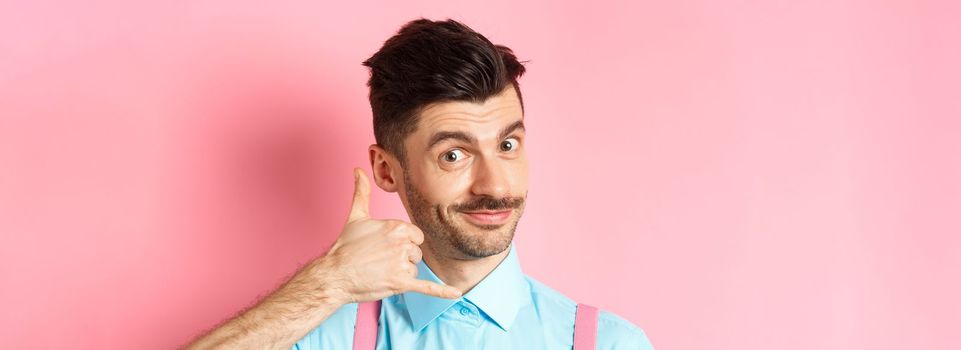 Close-up of handsome young guy with moustache, showing phone gesture, asking to call him, standing on pink background.
