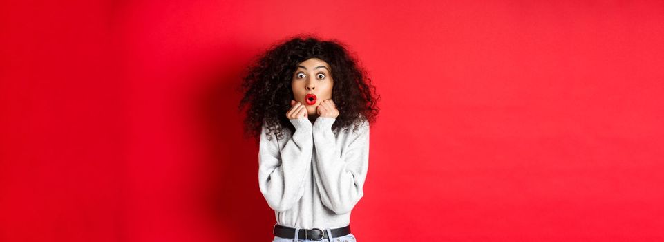 Surprised young woman looking with disbelief and amazement at camera, saying wow and standing in awe on red background.