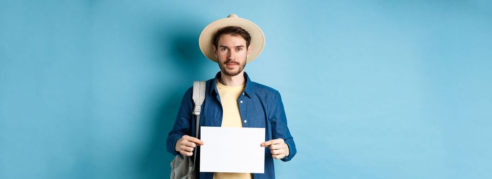 Image of smiling guy in straw hat backpacking, hitchhiking with piece of paper, travelling abroad on summer vacation, blue background.