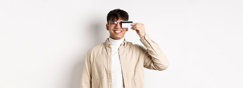 Cheerful young guy smiling real and showing plastic credit card, standing happy on white background, wearing glasses.